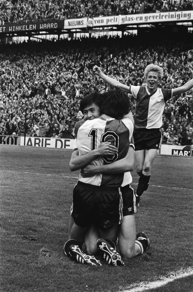 Drei Fußballspieler feiern ein Tor, indem sie sich auf dem Boden umarmen, vor einem jubelnden Publikum und schwenkenden Fahnen im Hintergrund, in Schwarz-Weiß aufgenommen.