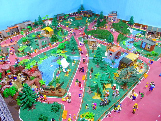 Ein Miniaturmodell eines Zoos mit Besuchern, umgeben von Bäumen, Häusern, Pflanzen, Gras, einem Zaun und anderen Gegenständen, vor einer Hintergrundwand mit Text unten.