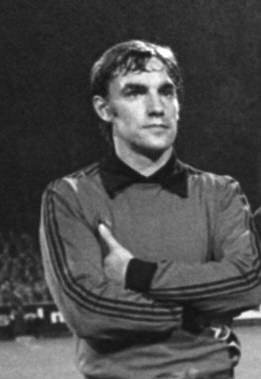 Schwarzes und weißes Foto eines Mannes, der mit verschränkten Armen auf einem Fußballfeld steht, umgeben von sitzenden Menschen im Hintergrund.