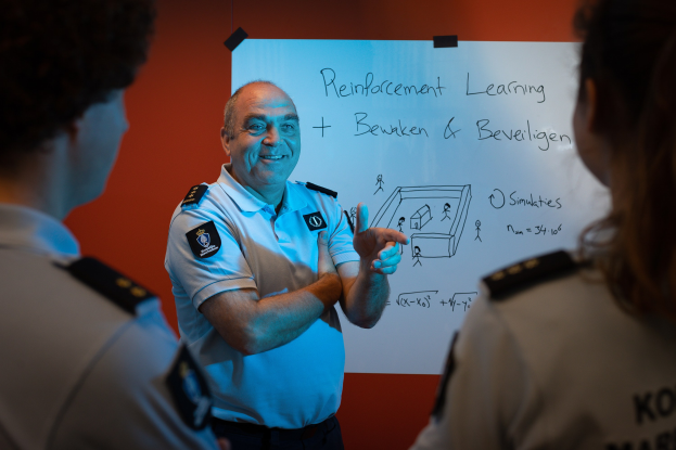 Ein Polizeibeamter in Uniform steht vor einer Whiteboard mit Text, flankiert von zwei anderen Individuen mit ernster Miene.