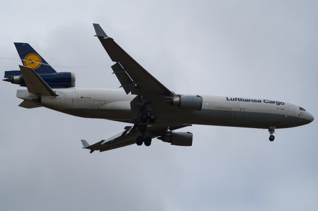 Ein Lufthansa Cargo Boeing 777-300ER im Flug, mit ausgebreiteten Flügeln und laufenden Triebwerken, sowie dem Text "Lufthansa Cargo" auf dem Rumpf gegen einen teilweise bewölkten Himmel.