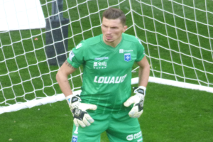 Ein Torwart in grüner Uniform steht vor einem Fußballtor auf saftig grünem Rasen, mit Handschuhen.