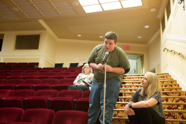 Ein Mann in einem grünen T-Shirt und blauen Jeans steht vor einem Publikum in einem Theater, hält ein Mikrofon in der Hand, zwei Frauen sitzen auf einer Treppe hinter ihm, rote Stühle im Hintergrund, ein Geländer auf der rechten Seite und Lampen auf dem Dach.