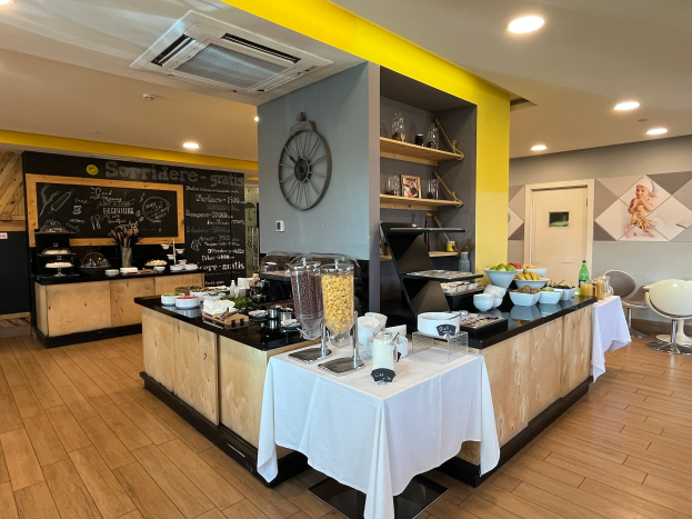 Ein Restaurant-Innenraum mit Tischen und Stühlen, Lebensmitteln, einer Tafel mit Text, einer Wanduhr, Deckenleuchten, einer Klimaanlage und einer Tür im Hintergrund, gelegen im Hôtel de Ville in Paris.