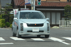 Ein silbernes Auto mit einer Person darin fährt eine Straße entlang neben einer Ampel, mit Gebäuden, Bäumen, Pflanzen, Geländern und einem Banner mit Text im Hintergrund.