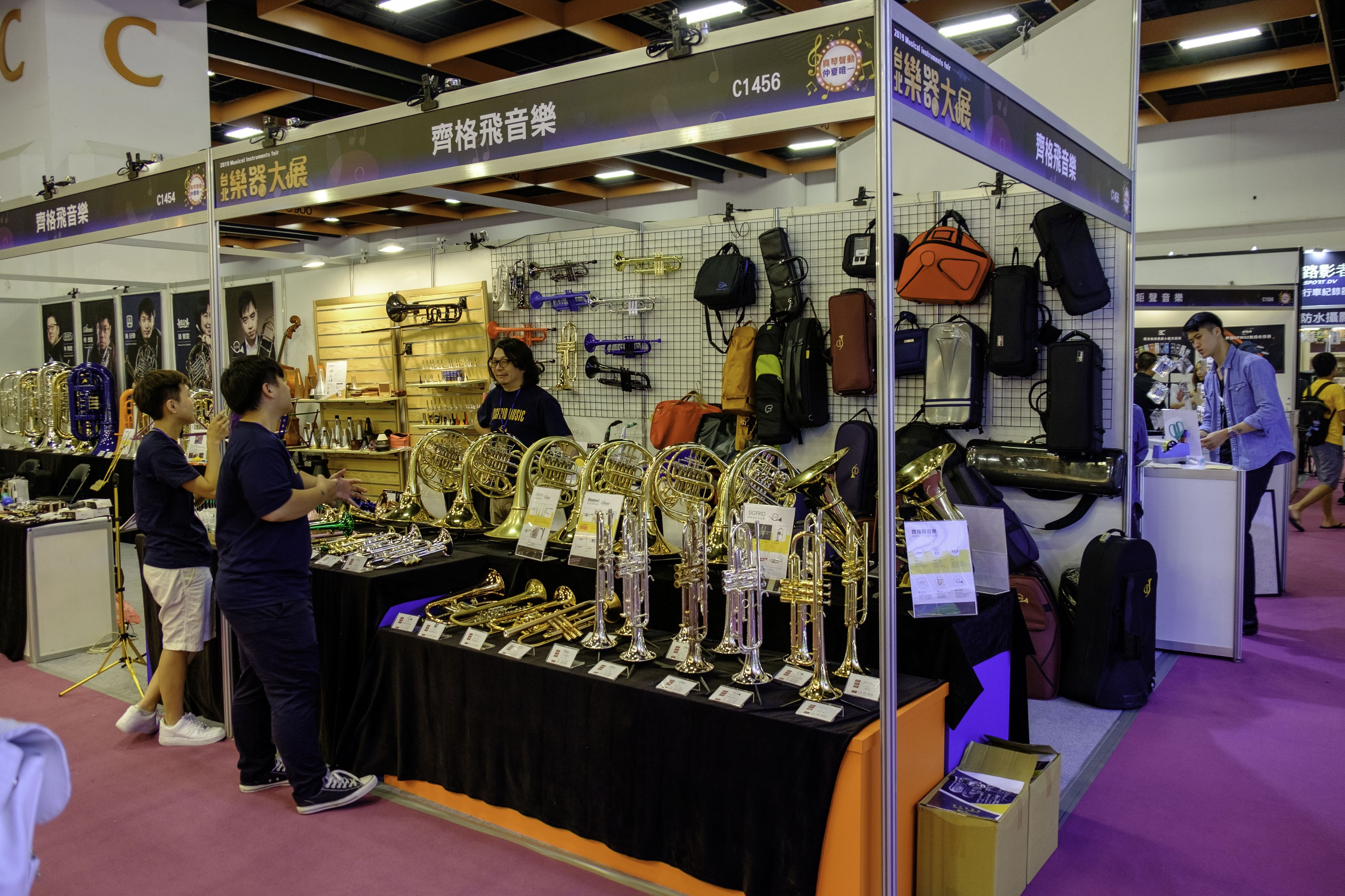 Eine Gruppe von Menschen um einen Tisch mit Musikinstrumenten, Tüten und Schildern mit Text, in einem Veranstaltungsort mit Deckenbeleuchtung und einem Karton auf dem Boden.