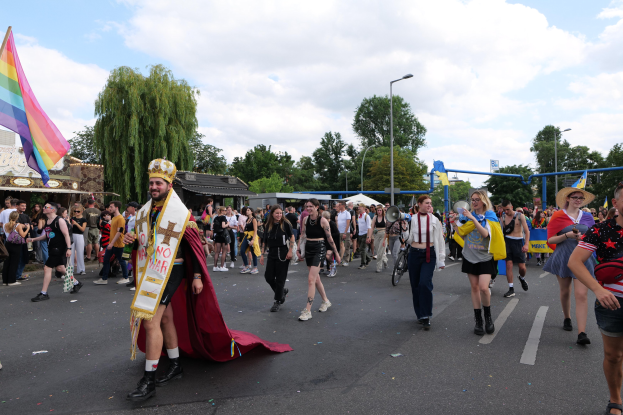 Menschen mit einer Regenbogenflagge und Musikinstrumenten auf der Straße während der Gay Pride Parade 2018, mit Bäumen, Schuppen und Laternen im Hintergrund.
