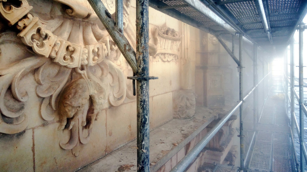 Gebäudefassade mit detaillierten Wandverzierungen links, Metallstangen rechts, Decke oben und Gerüst, das Renovierungsarbeiten anzeigt.