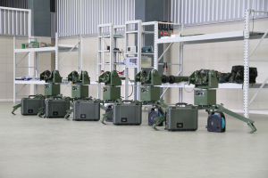 Gruppe von milit├Ąrischen Robotern auf einem Boden mit Koffern und Objekten auf Gestellen, gegen eine Wand mit Gel├Ąnder im Hintergrund.
