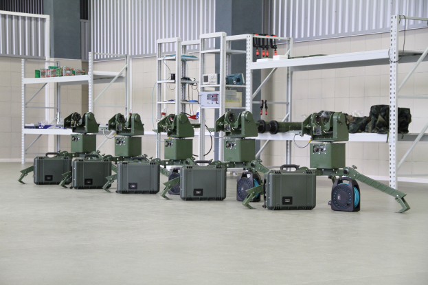 Gruppe von milit├Ąrischen Robotern auf einem Boden mit Koffern und Objekten auf Gestellen, gegen eine Wand mit Gel├Ąnder im Hintergrund.