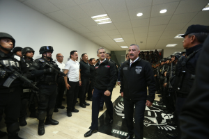 Eine Gruppe von Männern steht vor einer Reihe von bewaffneten, helmten Polizisten in einem Gerichtssaal, mit einer Wand und Deckenlampen im Hintergrund.