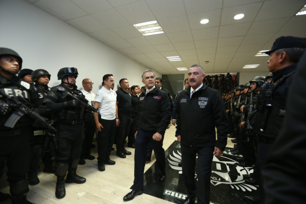 Eine Gruppe von Männern steht vor einer Reihe von bewaffneten, helmten Polizisten in einem Gerichtssaal, mit einer Wand und Deckenlampen im Hintergrund.