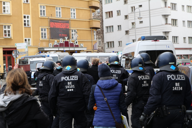 Polizei-Beamte in Uniform vor einer Menge bei einer Demonstration in Berlin, mit Fahrzeugen, Gebäuden und einem Kameramann im Bild.