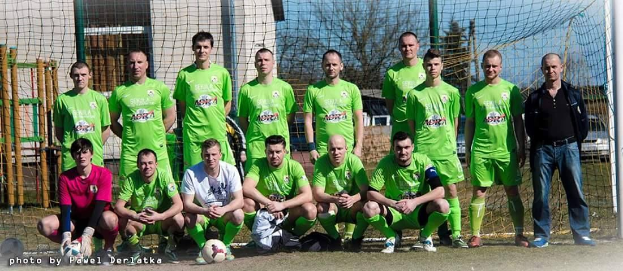 Gruppe von Männern in grünen T-Shirts, die auf einem Fußballfeld stehen und knien, mit einem Ball und einem Tor, Bäumen und Gebäuden im Hintergrund.