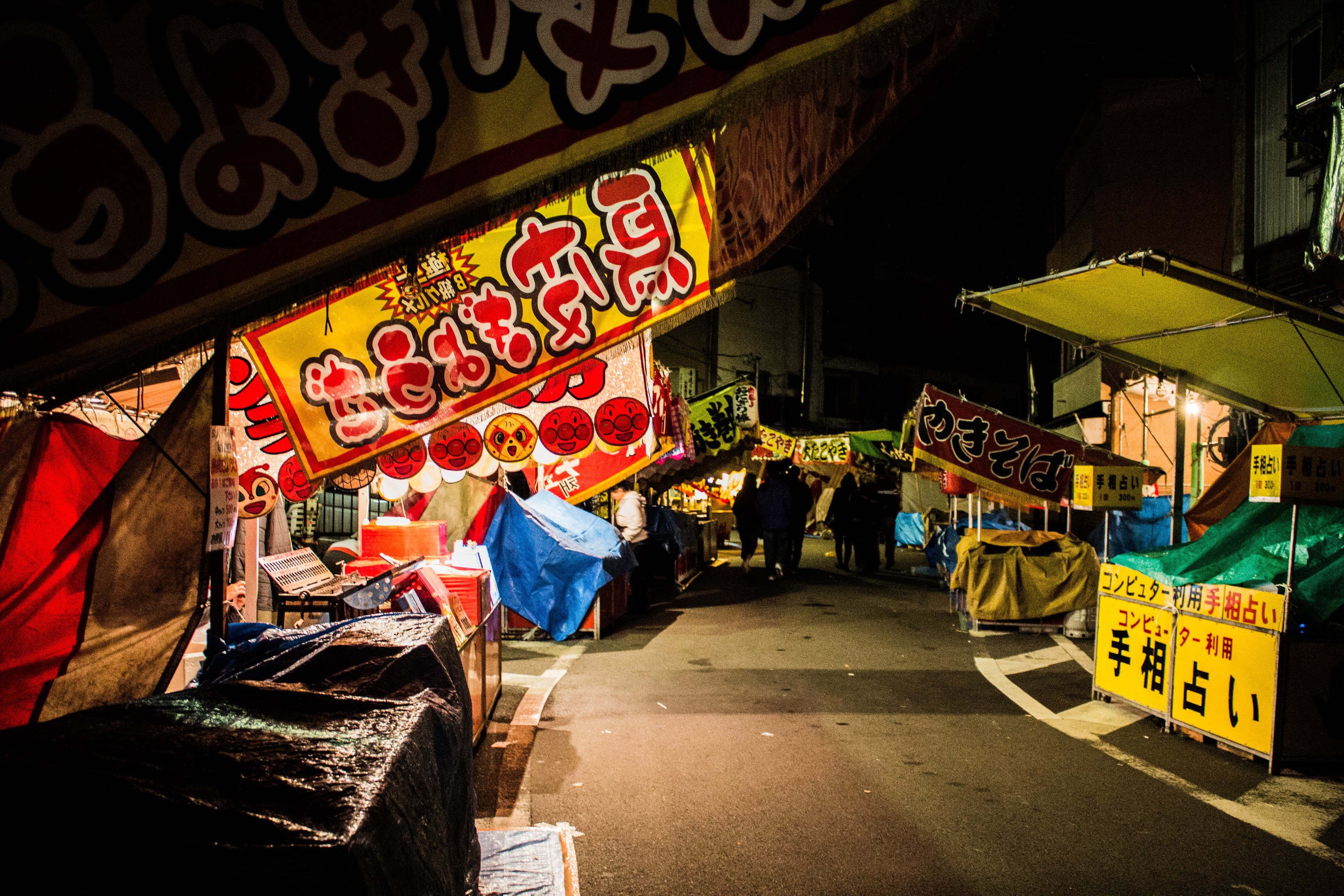 Ein belebter Nachtmarkt mit zahlreichen beleuchteten Ständen, die eine Straße säumen, Menschen, die zwischen ihnen umhergehen, und Gebäude auf beiden Seiten, mit einer Person, die zentral in der Mitte steht.