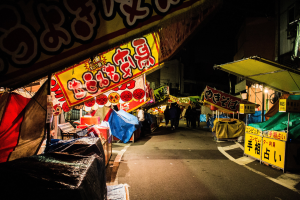 Ein belebter Nachtmarkt mit zahlreichen beleuchteten Ständen, die eine Straße säumen, Menschen, die zwischen ihnen umhergehen, und Gebäude auf beiden Seiten, mit einer Person, die zentral in der Mitte steht.