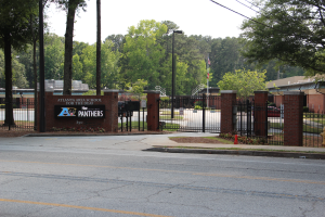 Der Eingang einer Schule mit einem Schild an der Seite, umgeben von Bäumen, Straßenlaternen, Gittern, Namensschildern, Fahrzeugen auf der Straße, Verkehrskegeln, Pflanzen, Gras, Gebäuden und einem klaren blauen Himmel.