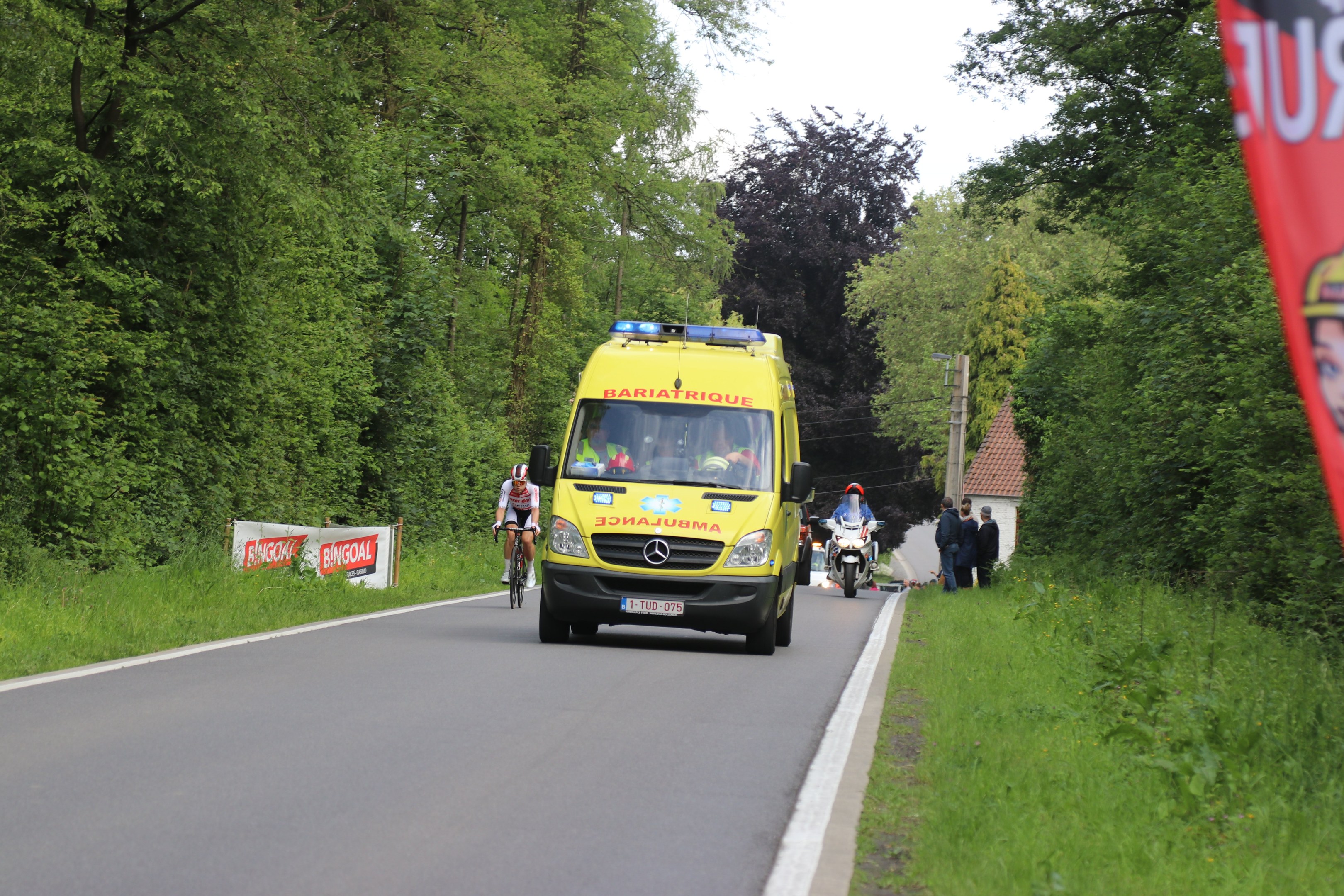 Ambulanz fährt auf einer Straße mit Radfahrern an den Seiten, umgeben von Gras, Bäumen, Häusern, Strommästen und einem klaren blauen Himmel.