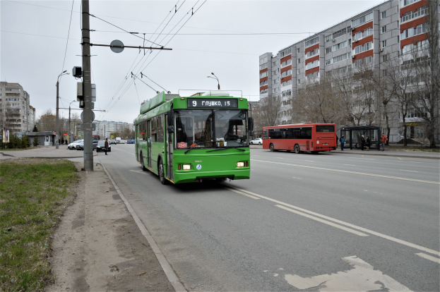 Ein grüner Bus fährt auf einer Straße mit hohen Gebäuden, Gras und Strommasten auf der linken Seite, Bäumen im Hintergrund und einem klaren blauen Himmel.