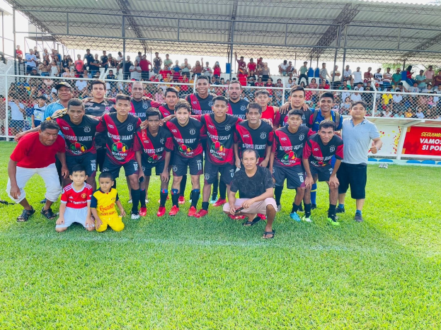 Gruppe von Männern, die auf einem Fußballfeld mit einem Siegesschild feiern, das 'Fotos Ugarte U-17 vs U-18' trägt, unter einem bewölkten Himmel.