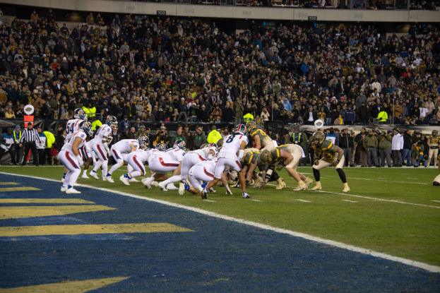 Ein Fußballspiel im Gange mit Spielern in Sportkleidung und Helmen auf dem Rasen, Stadionpublikum und Beschilderung im Hintergrund.