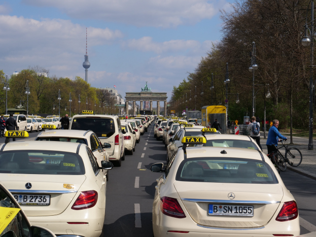 Eine lange Reihe von Taxis, die entlang einer belebten Straße in Berlin, Deutschland, geparkt sind, mit Fahrzeugen, Radfahrern und Fußgängern, flankiert von Laternen und Bäumen und Gebäuden, einem Bogen und einem Turm im Hintergrund unter einem bewölkten Himmel.