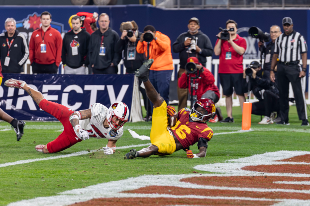 Ein Fußballspieler taucht in die Endzone für ein Touchdown ein, mit einer Gruppe von Menschen am Boden, einige halten Kameras, und ein Banner mit Text im Vordergrund, und Geländer, Pfosten und andere Gegenstände im Hintergrund.