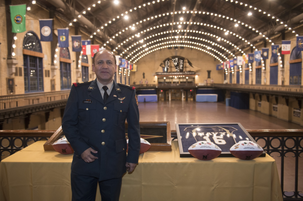 Ein Mann in Uniform, identifiziert als Stabschef der Nationalen Fußballliga (NFC), steht neben einem Tisch mit zwei Fußballen und einem Fotoframe, mit Flaggen und Lichtern im Hintergrund.