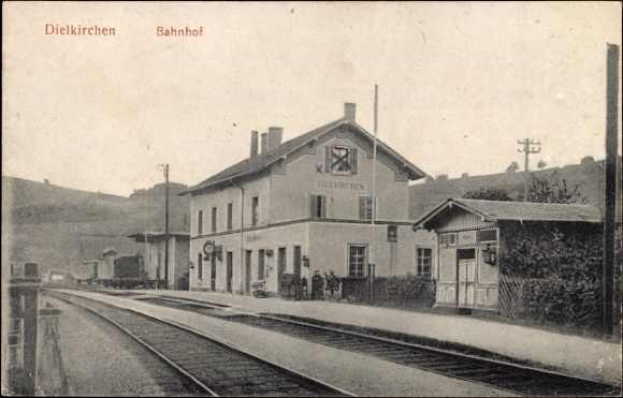 Schwarzes und weißes Foto einer Bahnstation in Dielkirchen, Deutschland, mit einem Zug auf den Gleisen, umgeben von Häusern, Strommasten, Bäumen, Menschen und Bergen sowie einem klaren Himmel.