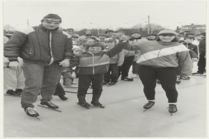Ein Schwarz-Weiß-Foto von einem Mann und einer Frau, die auf einem Eisstadion Schlittschuh laufen, umgeben von einer Gruppe von Menschen, einige tragen Jacken und Mützen, mit Bäumen, Pfählen, Gebäuden und einem klaren Himmel im Hintergrund.