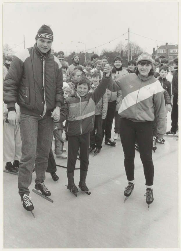 Ein Schwarz-Weiß-Foto von einem Mann und einer Frau, die auf einem Eisstadion Schlittschuh laufen, umgeben von einer Gruppe von Menschen, einige tragen Jacken und Mützen, mit Bäumen, Pfählen, Gebäuden und einem klaren Himmel im Hintergrund.