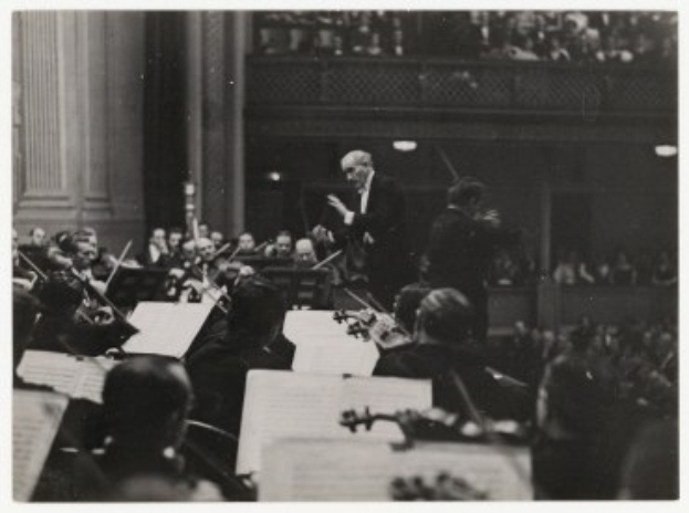 Ein Schwarz-Weiß-Foto eines Dirigenten vor einem Orchester, der einen Taktstock hält, mit sitzenden Musikern und Notenpulten im Vordergrund.