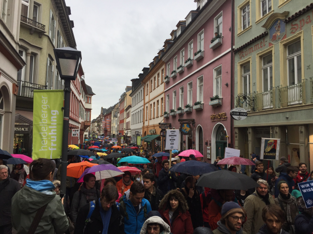 Große Gruppe von Menschen, die mit Schirmen die Straße entlanggehen, einige tragen Taschen und halten Schilder, mit Gebäuden auf beiden Seiten der Straße und einem Laternenmast im Vordergrund.