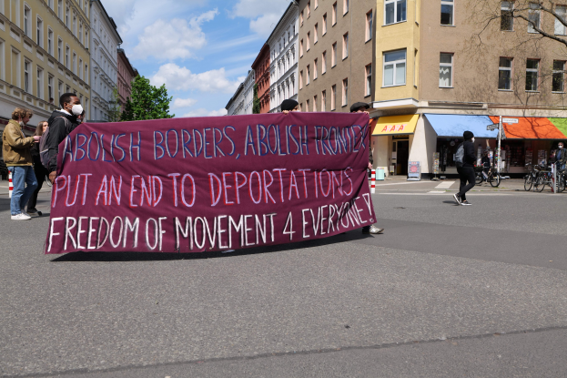 Eine Gruppe von Menschen marschiert auf einer Straße und hält ein Transparent mit der Aufschrift "Abolish Borders, Abolish Frontiers, Put an End to Deportations, Freedom of Movement 4 Everyone" hoch, mit Gebäuden, Bäumen und Fahrrädern, die die Straße säumen, unter einem bewölktem Himmel.