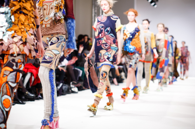 Eine Gruppe von Frauen, die auf dem London Fashion Week Laufsteg geht, mit einem Publikum im Hintergrund unter hellen Lichtern.