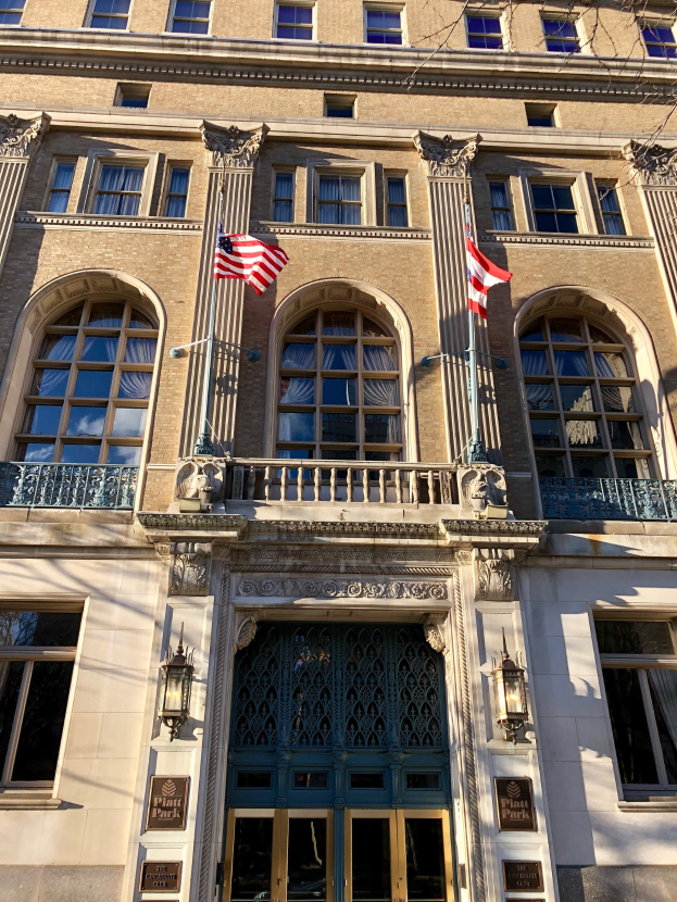 Außenansicht des Plaza Hotels in New York City, mit Glasfenstern, Türen, Säulen, Geländern, Leuchten, Schildern und Flaggen vor einem Himmel-Hintergrund.