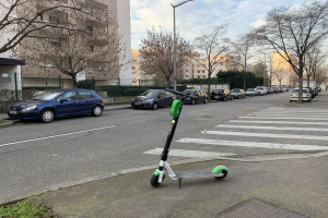 Ein elektrisches Scooter auf dem Straßenrand geparkt, mit Fahrzeugen, Laternenpfählen, Bäumen, Gebäuden mit Fenstern und einem bewölkten Himmel im Hintergrund.
