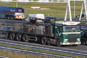Ein großes Frachtflugzeug mit Anhänger fährt auf einer Autobahn neben einer Brücke, mit Gras und Geländern auf beiden Seiten, unter einem klaren blauen Himmel mit grasigem Gelände und Masten im Hintergrund.