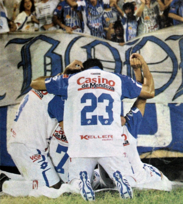 Gruppe von Fußballspielern, die auf dem Boden vor einer jubelnden Menge mit einem Banner im Hintergrund feiern.