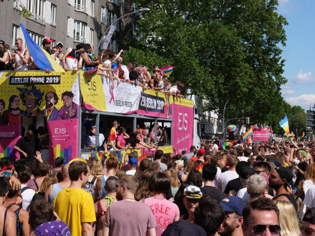 Eine große Menschenmenge, die während des Christopher Street Day 2019 in Berlin durch die Straßen geht, viele tragen Mützen und Schutzbrillen, halten Fahnen, mit Bannern und Gebäuden im Hintergrund bei bewölktem Himmel.