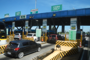 Eine Mautstation mit Autos davor, Fahrzeugen auf der Straße, Verkehrskegeln, Schildern mit Text, einer Brücke, Polen, Drähten, Bäumen und einem klaren blauen Himmel im Hintergrund.
