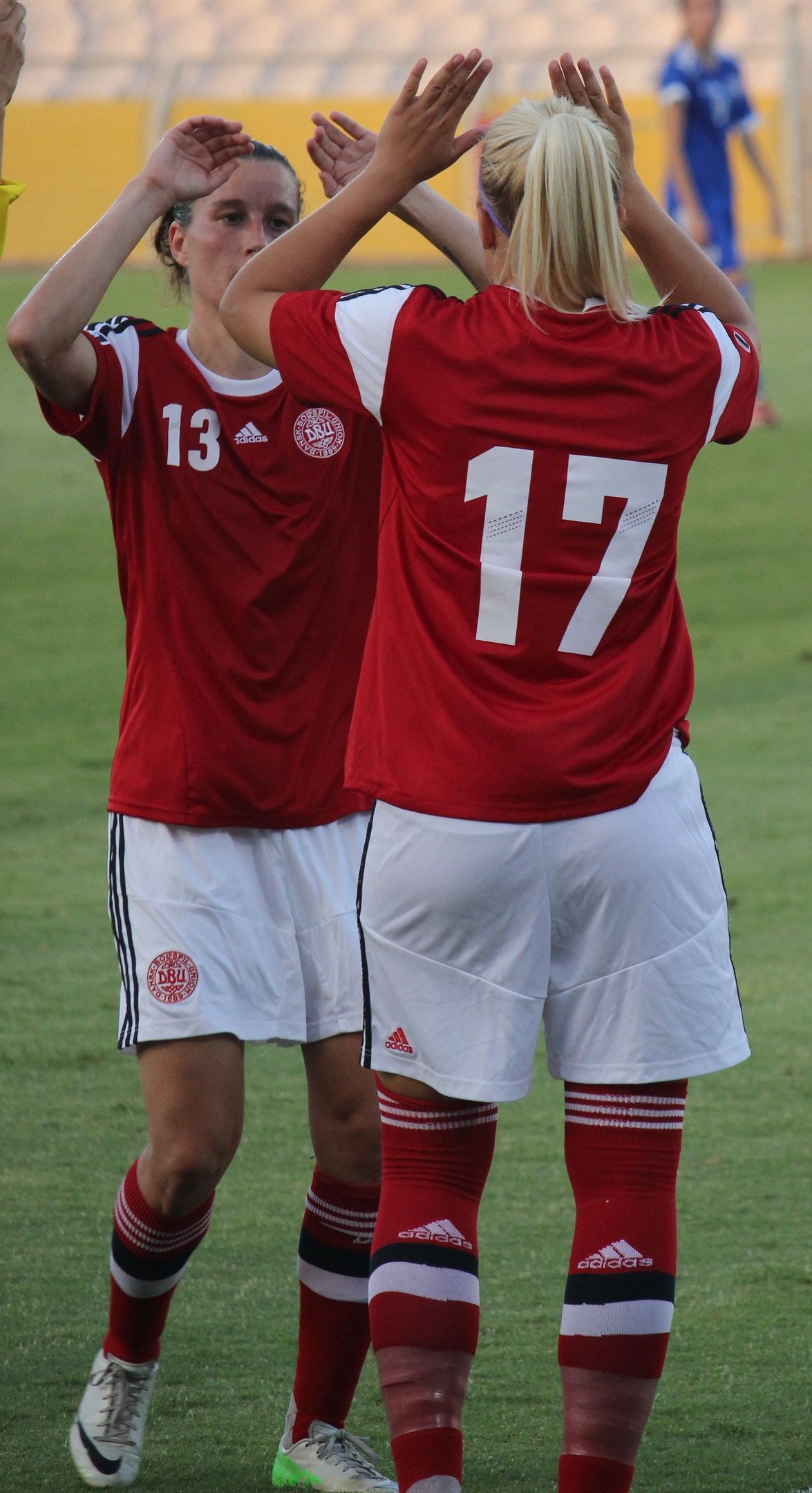 Zwei Frauen in roten T-Shirts und weißen Shorts klatschen auf einem Fußballfeld, mit einer Person in einem blauen Kleid und einem gelben Schild im Hintergrund.