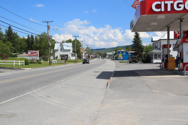 Eine Tankstelle am Straßenrand mit parkenden Autos, eine Tankstelle mit Namensschild und Fahne rechts und einem Hintergrund mit Bäumen, Häusern, Strommasten mit Drähten, einem Zaun, Gras, Pflanzen, Schildern mit Text und einem bewölkten Himmel.