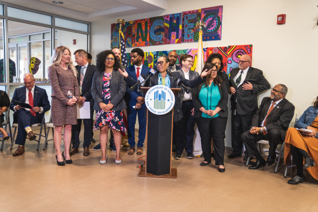 Gruppe von Menschen vor einem Rednerpult mit Mikrofon und einer Tafel mit der Aufschrift "Sancho Stadtrat tritt dem Stadtrat bei", mit sitzenden und stehenden Zuschauern, Fahnen, Feuerlöscher und Deckenleuchten im Hintergrund.