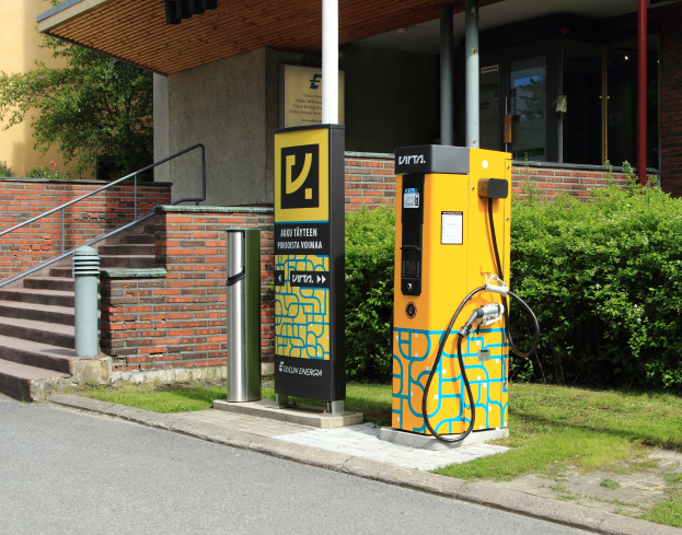 Eine Elektrofahrzeug-Ladestation an der Straße, umgeben von Grünflächen und einem Gebäude mit Fenstern und Säulen, mit Stufen und Geländern, die zu ihr führen.
