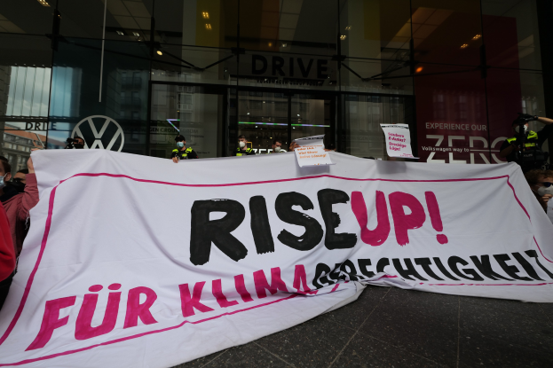 Eine Gruppe von Menschen mit Masken, die eine "Steh Auf"-Aufschrift und Schilder vor einem Glasgebäude halten, protestieren gegen eine Entscheidung der deutschen Regierung.