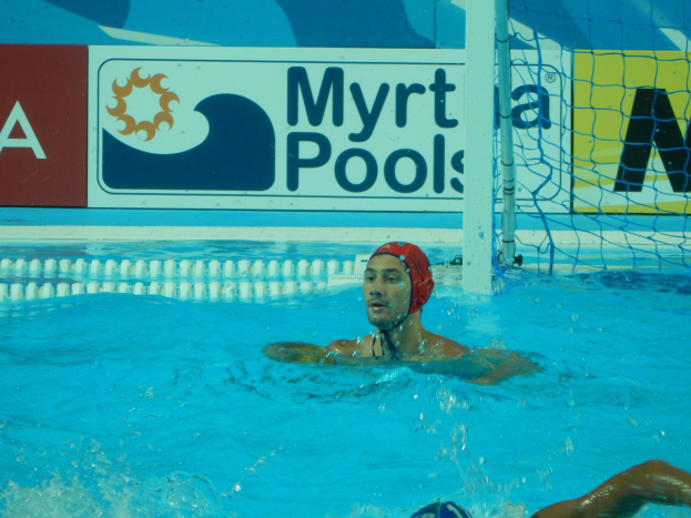 Wasserballspieler in einem Pool mit einer Mütze, der in der Nähe eines Netzes mit einer Werbetafel im Hintergrund schwimmt.