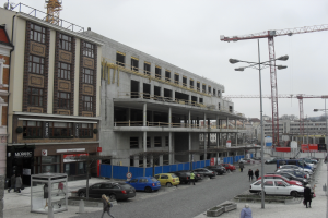 Ein großes Bauwerk im Bau in einer Stadt, umgeben von Straßenmasten, Straßenlaternen, Kraftfahrzeugen, Fußgängern, Infotafeln, Bäumen und einem klaren blauen Himmel.
