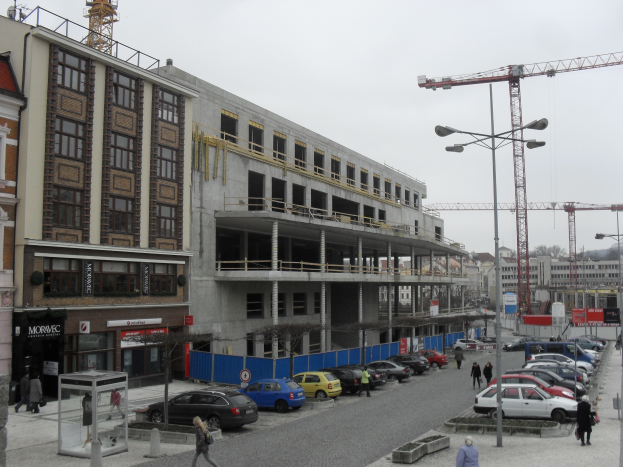 Ein großes Bauwerk im Bau in einer Stadt, umgeben von Straßenmasten, Straßenlaternen, Kraftfahrzeugen, Fußgängern, Infotafeln, Bäumen und einem klaren blauen Himmel.