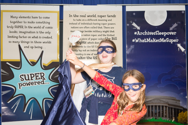 Zwei junge Mädchen in Superheldenkostümen posieren vor einem blauen Vorhang auf einem Schulereignis, eine hält ein Tuch, während beide Masken tragen.